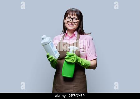 Frau mittleren Alters in Handschuhschürze mit organischen Reinigungsmitteln auf grauem Hintergrund Stockfoto