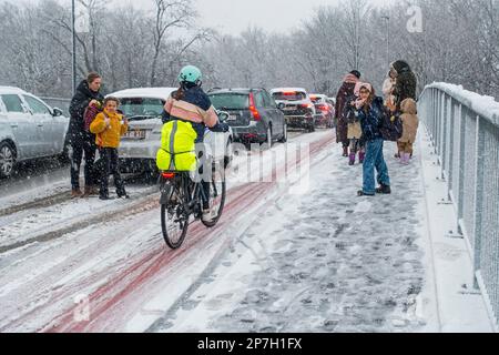 Radfahrer und Kinder, die auf rutschigem Pflaster auf einer Brücke zur Schule laufen, die während des unerwarteten späten Schneeschauers im März mit Schneeregen bedeckt war, Gent, Belgien Stockfoto