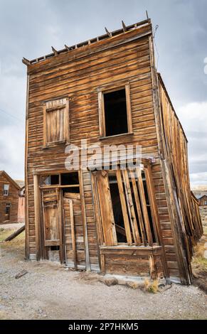 Ein hohes zweistöckiges Holzgebäude mit falscher Fassade lehnt an einer Seite, mit einer verbarrikadierten Tür und leeren Fenstern im Bodie State Historic Park, Kalifornien. Stockfoto
