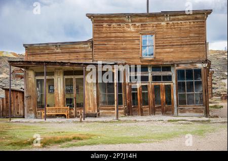 Ein zweistöckiges verwittertes Holzgebäude mit einer langen überdachten Veranda und geteilten Fenstern steht an einer Schotterstraße im Bodie State Historic Park, Kalifornien. Stockfoto