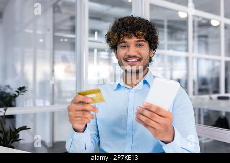 Ein junger spanischer Mann sitzt im Büro, hält ein Telefon und benutzt eine Kreditkarte. Ein Lächeln, das auf die Kamera zeigt. Stockfoto