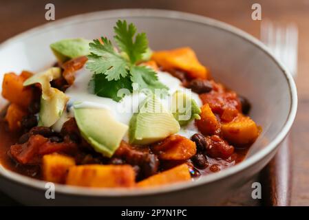 Schüssel mit einer hausgemachten veganen Süßkartoffel-Chili aus schwarzen Bohnen mit Sojaboghurt und Avocado-Stücken. Stockfoto