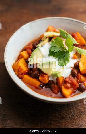 Schüssel mit einer hausgemachten veganen Süßkartoffel-Chili aus schwarzen Bohnen mit Sojaboghurt und Avocado-Stücken. Stockfoto