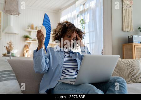 Schwarze Frau leidet unter Hitzschlag, flach ohne Klimaanlage, die den Ventilator winkt, wenn sie zu Hause auf der Couch sitzt Stockfoto