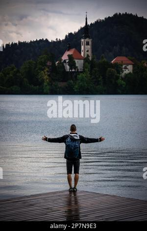 Junger weißer Mann, der mit den Händen auseinander steht und die Umgebung auf einem hölzernen Pier umarmt, während er die berühmte Kirche in Bled, Slowenien, beobachtet Stockfoto