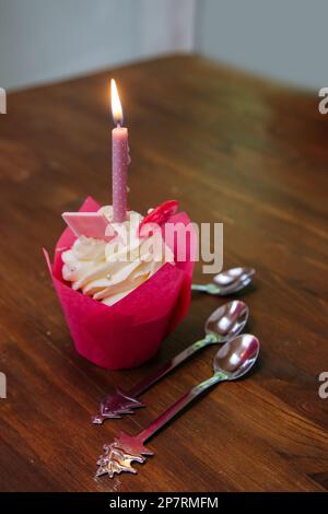 Leckerer Cupcake für die Geburtstagsparty mit Kerze und Löffeln auf einem braunen Holztisch. Kuchen mit weißer Käsesahne, pinkfarbenen Lutscher und Schokolade. Stockfoto