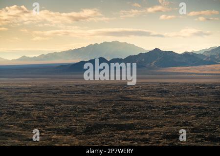 Weiches Morgenlicht fällt über eine weite dunkle Lavaebene mit verschwommenen Bergkämmen, gesehen vom Amboy Crater, Mojave Trails National Monument, Kalifornien. Stockfoto