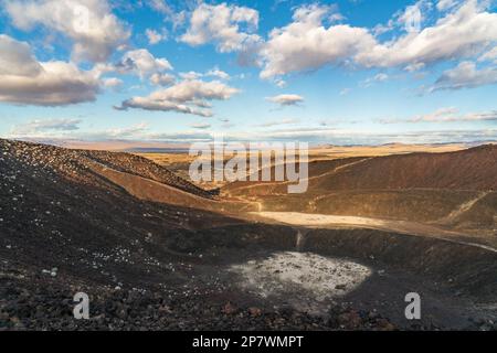 Ein kleiner entfernter Wanderer steht auf dunklem Lavagestein am Rand eines weiten hellen Wüstentals am Amboy-Krater, Mojave Trails National Monument, Kalifornien. Stockfoto