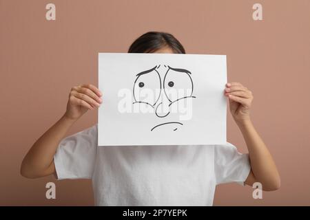 Ein kleines Mädchen versteckt sich hinter einem Blatt Papier mit traurigem Gesicht auf blassrosa Hintergrund Stockfoto