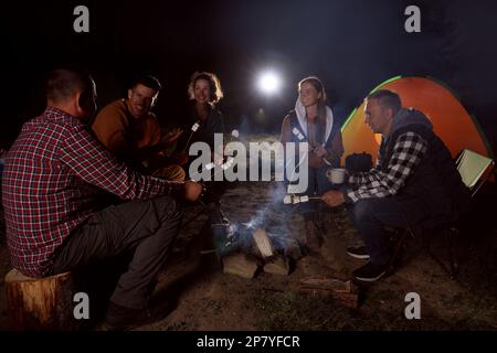 Eine Gruppe von Freunden, die abends am Lagerfeuer auf dem Campingplatz Marshmallows rösten Stockfoto