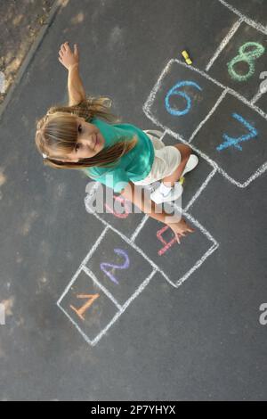 Kleines Mädchen, das Hopscotch spielt, gezeichnet mit Kreide auf Asphalt draußen, über der Aussicht. Glückliche Kindheit Stockfoto