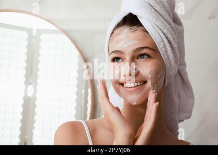 Ein wunderschönes Teenager-Mädchen trägt Reinigungsschaum auf das Gesicht im Badezimmer auf, Platz für Text. Hautpflegekosmetik Stockfoto