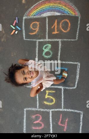 Kleines afroamerikanisches Mädchen und farbenfroher Hopscotch, gezeichnet mit Kreide auf Asphalt, Blick von oben. Glückliche Kindheit Stockfoto