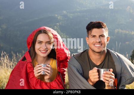 Glückliches Paar mit Getränken in Schlafsäcken umgeben von wunderschöner Natur Stockfoto