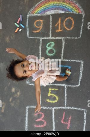 Kleines afroamerikanisches Mädchen und farbenfroher Hopscotch, gezeichnet mit Kreide auf Asphalt, Blick von oben. Glückliche Kindheit Stockfoto