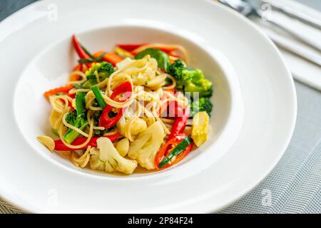 Pad Kee Mao oder thailändische Fusionsküche, gebratene Spaghetti mit scharfen Kräutern, Fleisch und frischem Gemüse. Schönes thailändisches Essen in einem weißen Gericht auf einem Tisch, Stockfoto