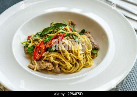 Köstliche thailändische Fusionsküche, gebratene Spaghetti mit thailändischem grünem Curry und Fleisch. Wunderschönes thailändisches Essen in einem modernen weißen Gericht auf einem Tisch im Restaurant, Stockfoto