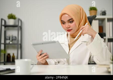 Professionelle und konzentrierte asiatische muslimische Geschäftsfrau oder Büroangestellte, die Hijab mit ihrem digitalen Tablet trägt und sich auf ihren Bu konzentriert Stockfoto