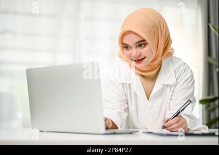 Attraktive und selbstbewusste muslimische Geschäftsfrau der Millennials, die sich auf den Tisch lehnt, auf den Laptop-Bildschirm schaut, etwas auf dem Bericht notiert, in ihrem Büro arbeitet Stockfoto