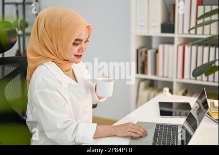 Eine wunderschöne und selbstbewusste asiatische muslimische Geschäftsfrau oder CEO, die ihren Morgenkaffee trinkt und ihr Notebook an ihrem Schreibtisch benutzt Stockfoto