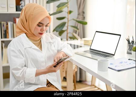 Professionelle und selbstbewusste asiatische Geschäftsfrau, die Hijab trägt und ihr Telefon an ihrem Schreibtisch in einem modernen Privatbüro benutzt. Stockfoto