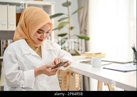 Wunderschöne junge asiatische muslimische Geschäftsfrau oder Unternehmerin, die Hijab und Freizeitkleidung trägt und ihr Telefon im Büro benutzt. Stockfoto