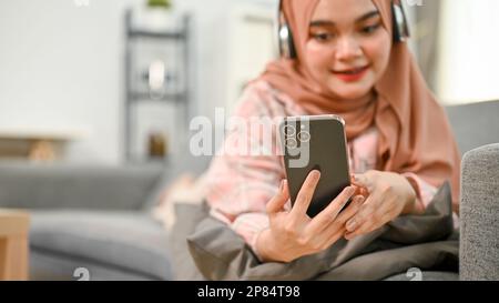 Glückliche junge asiatische Muslime in lässigen Kleidern und Hijab, die Musik über Kopfhörer hören, ihr Telefon benutzen, auf dem Sofa in ihrem Wohnzimmer liegen. Stockfoto