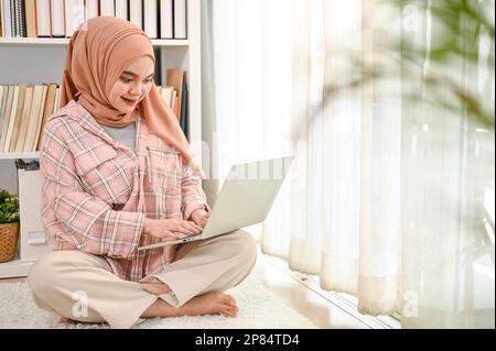 Wunderschöne junge asiatische Muslime, die Hijab trug, ihren Laptop in ihrem Wohnzimmer benutzte und von zu Hause aus arbeitete. Stockfoto