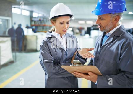 Optimierung der Versandlogistik in der Fabrik. Zwei Manager in Arbeitskleidung, die sich über ein Klemmbrett unterhalten, während sie auf dem Druck stehen Stockfoto