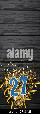 Blaue Festkerze Nummer 27 und goldene Konfetti auf dunklem Holzhintergrund. 27. Geburtstagskarte. Jubiläums- und Geburtstags Vertikales Banner. Stockfoto