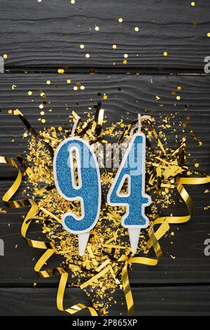 Blaue Festkerze Nummer 94 und goldene Konfetti auf dunklem Holzhintergrund. 94. Geburtstagskarte. Jubiläums- und Geburtstags Vertikales Banner. Stockfoto