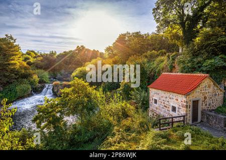 Krka, Kroatien - wunderschöne Sommersonnenaufgangslandschaft eines kleinen Steinhauses an den Krka-Wasserfällen im Krka-Nationalpark, Dalmatien, mit warmen Morgenfarben Stockfoto