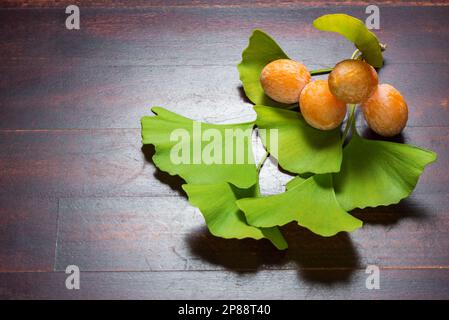 Ginkgo biloba reife gelbe Früchte und grüne Blätter auf Holzhintergrund. Stockfoto