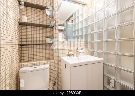 Stilvolles und einfaches Badezimmerdesign mit beigefarbenem Mosaik und Toilette sowie Glasblockwand mit Regalen. Konzept-Hotelzimmer oder Designerarbeit. Europäisch Stockfoto