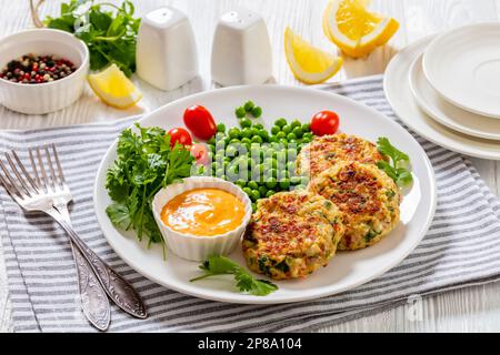 Krabbenkuchen mit Remouladesauce, grünen Erbsen, Tomaten, frischem Koriander auf weißem Teller auf weißem Holztisch, horizontale Ansicht Stockfoto