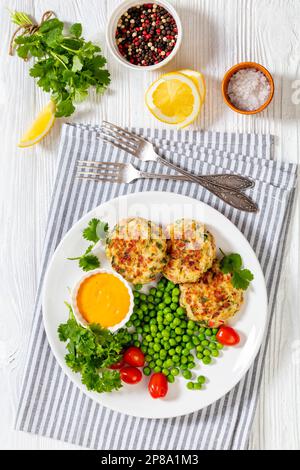 Krabbenkuchen mit Remouladesauce, grünen Erbsen, Tomaten, frischem Koriander auf weißem Teller auf weißem Holztisch, vertikale Ansicht von oben Stockfoto