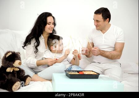 Familie in weißen Kleidern, die mit Essstäbchen aus orientalischem Essen spielt Holzstäbchen Trommel Lachen viel Spaß im Bett zusammen essen Sushi weiches Spielzeug brauner Teddybär Tag frei Papa mama Sohn Stockfoto