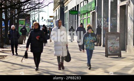 Warschau, Polen. 8. März 2023 Eine Menge Leute, die einen urbanen Bürgersteig entlang laufen, mit hellem, glühendem Sonnenlicht im Hintergrund auf einer belebten Straße in Down to Stockfoto