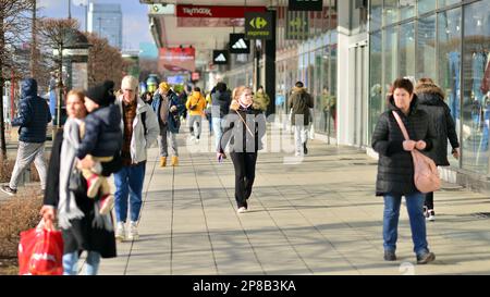 Warschau, Polen. 8. März 2023 Eine Menge Leute, die einen urbanen Bürgersteig entlang laufen, mit hellem, glühendem Sonnenlicht im Hintergrund auf einer belebten Straße in Down to Stockfoto