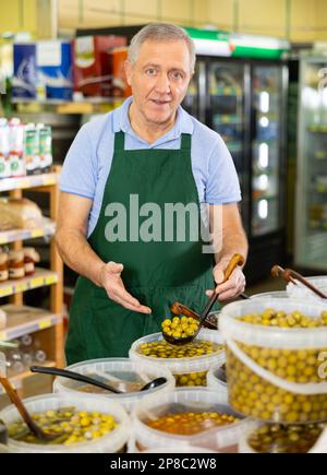 Positiver alter männlicher Händler in der Schürze, der Oliven mit einer Schaufel aus einem Kunststoffbehälter im Lebensmittelgeschäft aufnimmt Stockfoto