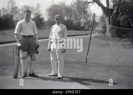 Etwa 1930er, historisch, ein junger Mann mit Polstern, Handschuhen und einem Schläger, der ein Cricketnetz auf dem Rasen eines Landhauses in England hält. Er steht neben einem älteren erwachsenen Cricketspieler, dem Bowler, der das Cricket-Weiß des Tages trägt, Hosen oder Flanelle, die von einer Krawatte, einem Rundhalsausschnitt-Hemd und einer Kappe getragen werden. Stockfoto