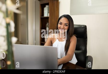 Eine fröhliche junge indische Geschäftsfrau sitzt in ihrem Büro und arbeitet mit einem Notebook, das drahtlose Technologie nutzt und über das Internet kommuniziert. Stockfoto