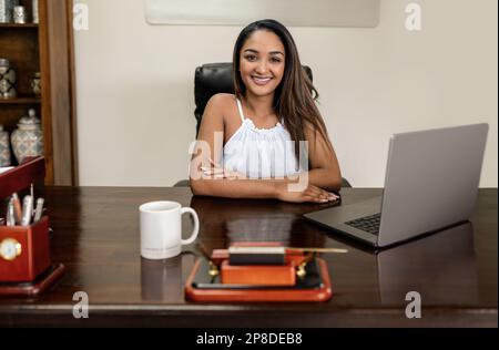 Eine fröhliche, junge, Erwachsene Anwältin aus Indien sitzt an einem Tisch und nutzt ihren Laptop und drahtlose Technologie für die Geschäftskommunikation, während sie in den lächelt Stockfoto