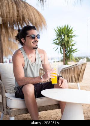 Lateinamerikanischer Mann mit langen schwarzen Haaren, der ein Orangengetränk auf der Terrasse der Strandbar genießt Stockfoto