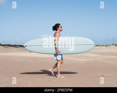 Hispanischer Surfer, der auf dem Sand mit seinem Surfbrett spaziert Stockfoto