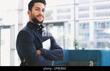 Selbstbewusst und stolz. Verkürztes Porträt eines gutaussehenden jungen Geschäftsmanns, der im Büro mit gefalteten Armen und einem Tablet in der Hand steht. Stockfoto
