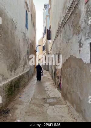 Rückansicht einer Frau in einem traditionellen schwarzen Djellaba-Bademantel, die eine enge Gasse in Essaouira, Marokko, entlanggeht Stockfoto