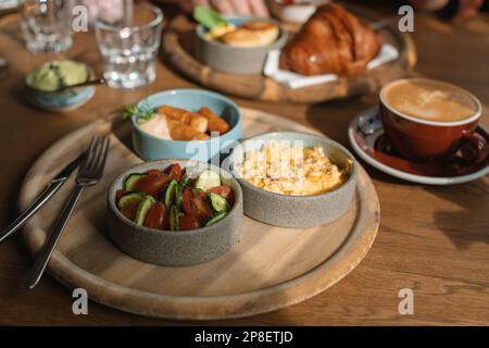 Frühstückstisch mit Rührei, Würstchen, Avocado, Croissant, Gurke, Tomaten, Kaffee und Wasser Stockfoto