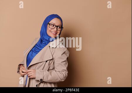 Stilvolle, elegante, selbstbewusste muslimische Frau in blauem Hijab und Brille, lächelnd auf isolierten beigen Hintergrund Stockfoto