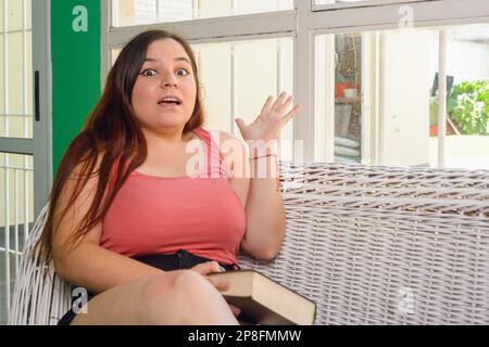 Junge argentinische Latina-Frau, die zu Hause auf der Couch saß und beim Lernen überrascht war, in die Kamera schaute, mit offenem Mund und die Hand machte Stockfoto